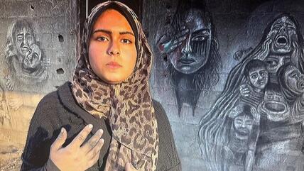 A woman in an animal-print headscarf and black jumper (Menna Hamouda) stands in front of charcoal and chalk drawings on a wall in Gaza; she looks very serious