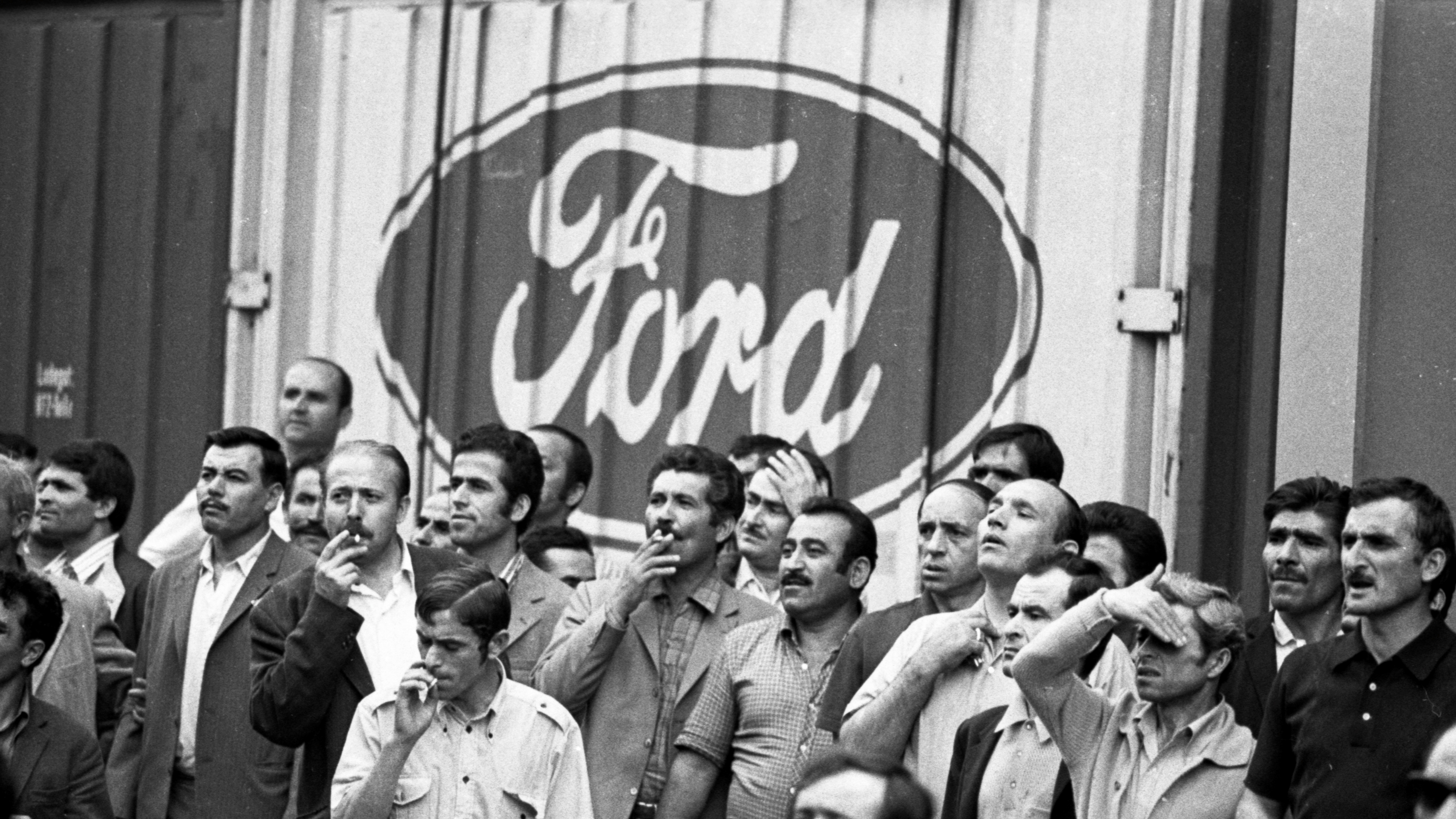 Streikende 1973 im Ford-Werk Köln. Foto: Picture Alliance | Klaus Rose Schwarz-weiß Bild von Männern, die vor einer Wand stehen. Auf der Wand ist das Firmenlogo von Ford zu sehen.