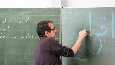 Islamic religious education in Bonn, 2022. (Photo: Picture Alliance / KNA | H. Oppitz) A man wearing glasses writes in Arabic on a green board.